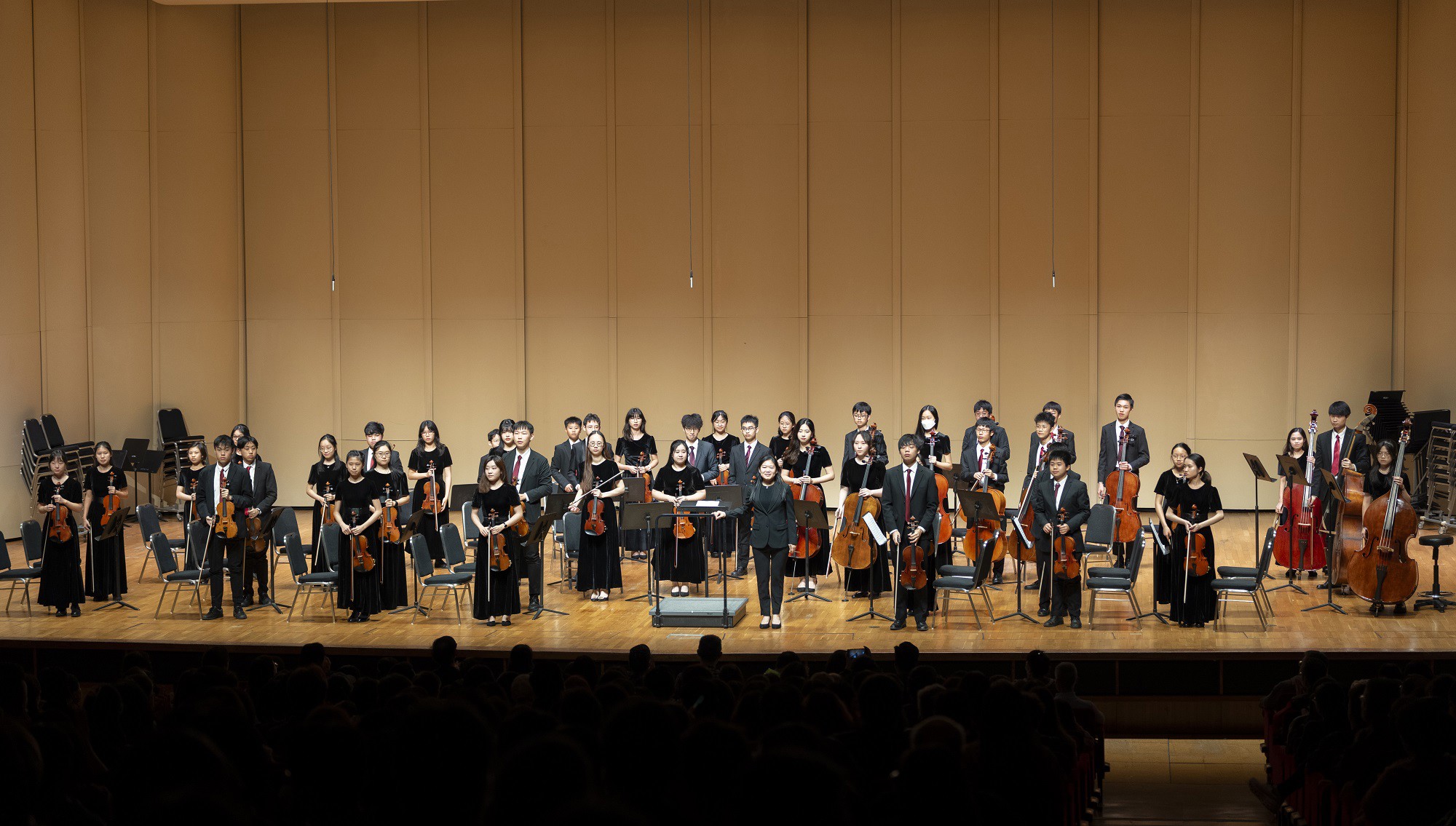 Orchestra performing on stage with conductor leading musicians in formal black attire with string instruments.