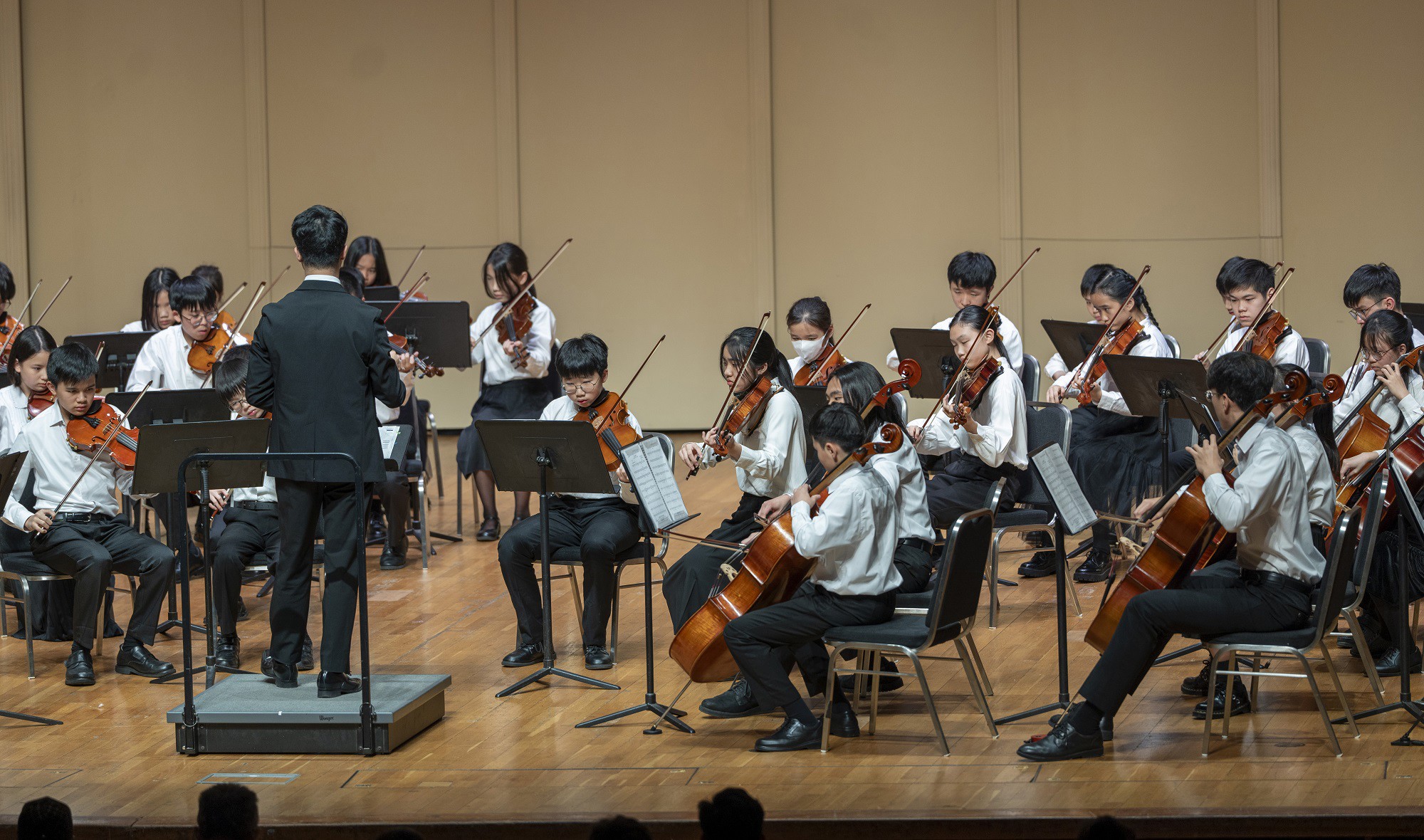 Student orchestra performing with conductor leading young musicians playing string instruments in concert hall.